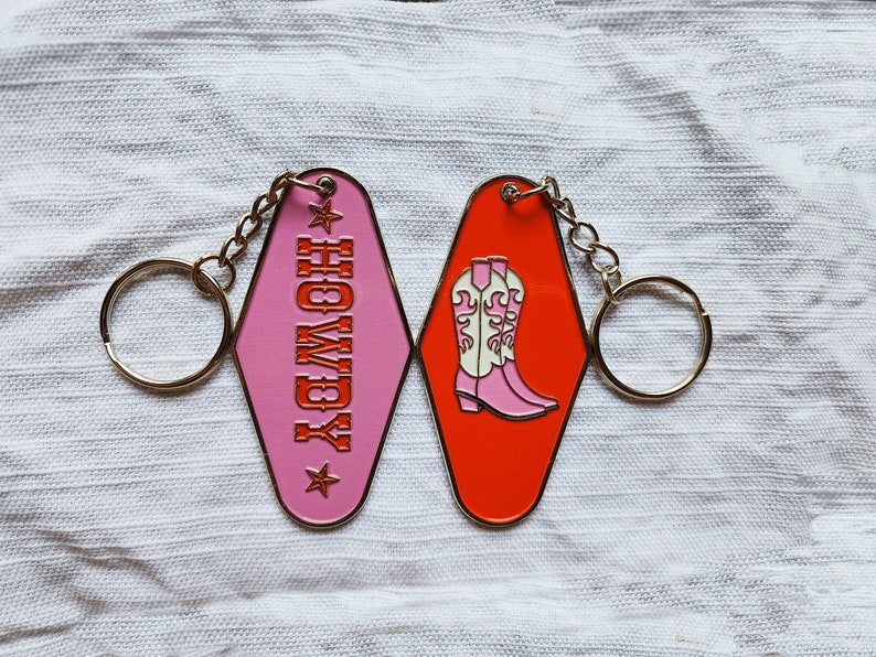 May include: Pink and orange motel keychains with a gold chain and ring. The pink keychain says "Howdy" with a star on each side. The orange keychain has a pair of pink cowboy boots with white tops.