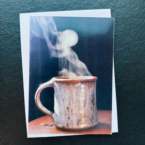 May include: A greeting card featuring a ceramic mug with steam rising from it. The mug is brown with a speckled design and a reddish-brown rim. The background is dark blue, and the card is on a white background.