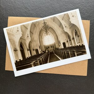 May include: A sepia-toned photograph of a church interior, featuring rows of pews, arched ceilings, and a large chandelier. The image is on a white card, resting on a brown envelope, set against a dark background. The church's architecture is the focal point.