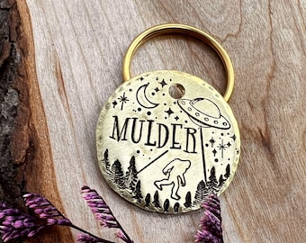 May include: A round brass pet tag with a gold-toned split ring featuring an engraved design of a UFO beaming down a Bigfoot figure, a crescent moon, stars, and pine trees, with the name MULDER centered in bold, stylized black lettering.