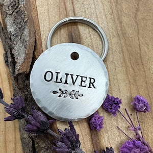 Könnte beinhalten: Ein runder, silberner Haustieranhänger mit dem Namen "OLIVER" in schwarzer Schrift und einem dekorativen Blattzweig. Der Anhänger ist an einem silbernen Schlüsselring befestigt. Lila Lavendelzweige und -knospen sind um den Anhänger herum angeordnet.