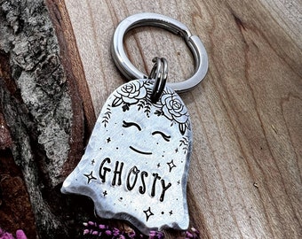 Può includere: Un portachiavi in metallo argentato a forma di fantasma con una texture martellata. Il fantasma presenta una corona floreale incisa, occhi chiusi, una bocca semplice e la parola GHOSTY circondata da piccoli accenti a stella e punti, su un anello.