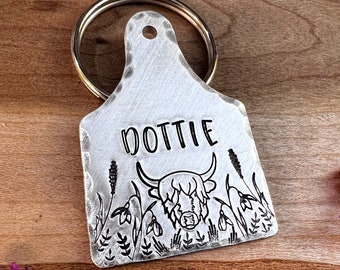 May include: Silver metal keychain with a cow silhouette and the name "Dottie" engraved on it. The cow is surrounded by flowers and grass.