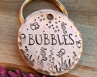 May include: Copper dog tag with a gold key ring. The tag has a hammered texture and features an engraved design of seahorses, jellyfish, and bubbles. The text "BUBBLES" is engraved in the centre of the tag.