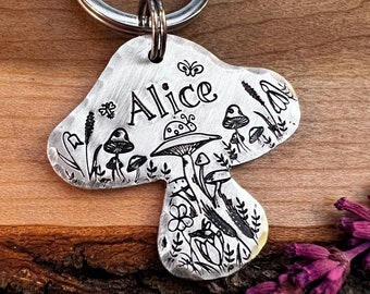 Può includere: Un'etichetta per cani in argento a forma di fungo con il nome "Alice" inciso su di essa. L'etichetta presenta un design dettagliato di funghi, fiori e una farfalla.