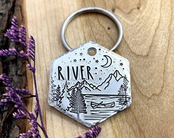 May include: Silver keyring with a hexagonal charm featuring a mountain scene with a canoe on a lake, stars, a crescent moon, and the word "RIVER" engraved on it.