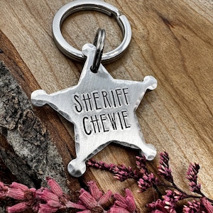 Op de afbeelding: Een zilveren sheriffster-sleutelhanger met de tekst "SHERIFF CHEWIE" erop gegraveerd. De sleutelhanger is bevestigd aan een zilveren sleutelring en ligt op een houten oppervlak met roze bloemen.