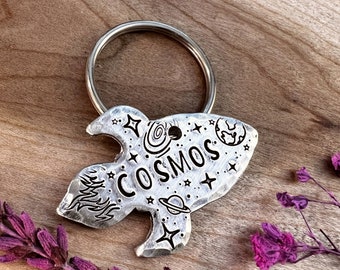 May include: Silver rocket ship keychain with the word "COSMOS" engraved on it. The keychain features a small planet, stars, and a swirl design.