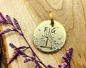 Puede incluir: Una etiqueta redonda para mascotas de metal dorado, con un diseño de velero y olas. La etiqueta está grabada con el nombre "FIG" y tiene un pequeño anillo para sujetarla a un collar.