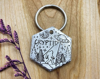 May include: A hexagonal silver-toned metal keychain featuring a hammered edge and an engraved design of a Bigfoot figure, a UFO, a moon, and pine trees. The word CRYPTID is etched in black lettering across the center of the metal surface.