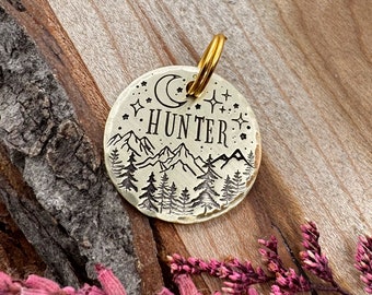 May include: A brass dog tag with a mountain scene, a crescent moon, and stars. The tag is engraved with the name "HUNTER".