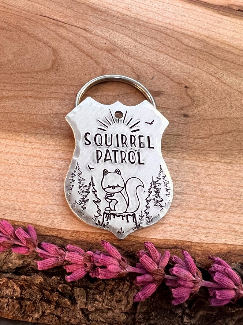 May include: A silver metal keychain shaped like a badge with the text "Squirrel Patrol" and a cartoon squirrel sitting on a tree stump. The keychain has a ring attached to the top.
