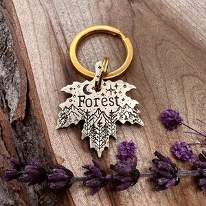 Könnte beinhalten: Ein metallener schlüsselförmiger Anhänger in Blattform mit einem goldenen Ring. Der Blattanhänger zeigt ein Berg- und Waldmotiv mit dem Wort "Forest" sowie Mond und Sterne. Der Schlüsselanhänger liegt auf einer Holzoberfläche mit lila Blumen.