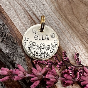 May include: A round brass pet tag featuring a floral design and the name "Ella" engraved on it. The tag has a gold-coloured ring for attaching to a collar.