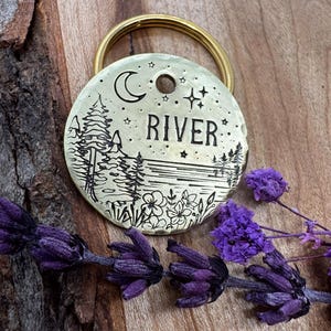 K&ouml;nnte beinhalten: Ein Messing-Hundeanh&auml;nger mit einer eingravierten Mond, Sternen und einer Waldszene. Der Anh&auml;nger tr&auml;gt den Namen "RIVER".