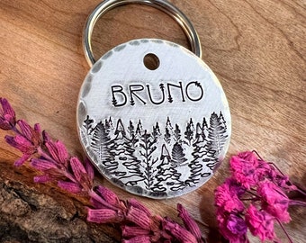 May include: A silver round dog tag with a forest scene engraved on it. The tag has a ring attached and the name "BRUNO" is engraved on the top.