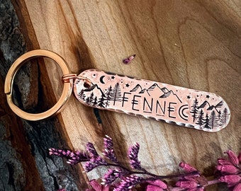 May include: A copper-colored keychain with a mountain scene and the word "FENNEC" engraved on it. The keychain is on a wooden surface with pink flowers.