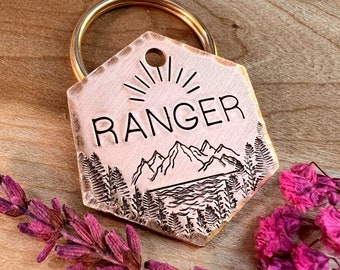Könnte beinhalten: Ein sechseckiger Schlüsselanhänger aus Kupfer mit einem goldenen Ring. Der Schlüsselanhänger ist mit einem eingravierten Design einer Bergkette, eines Sees und einer Sonne mit dem Wort "RANGER" über den Bergen verziert.