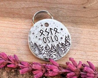 May include: Silver dog tag with a hammered finish, featuring a forest scene with ferns and the word "OSLO" in a stylized font. The tag also includes a runic symbol and stars.