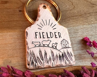 May include: Copper-colored metal tag with the word "FIELDER" and a farm scene etched on it. The scene includes silhouettes of cows, sheep, a barn, and wheat. A gold-colored ring is attached to the tag.
