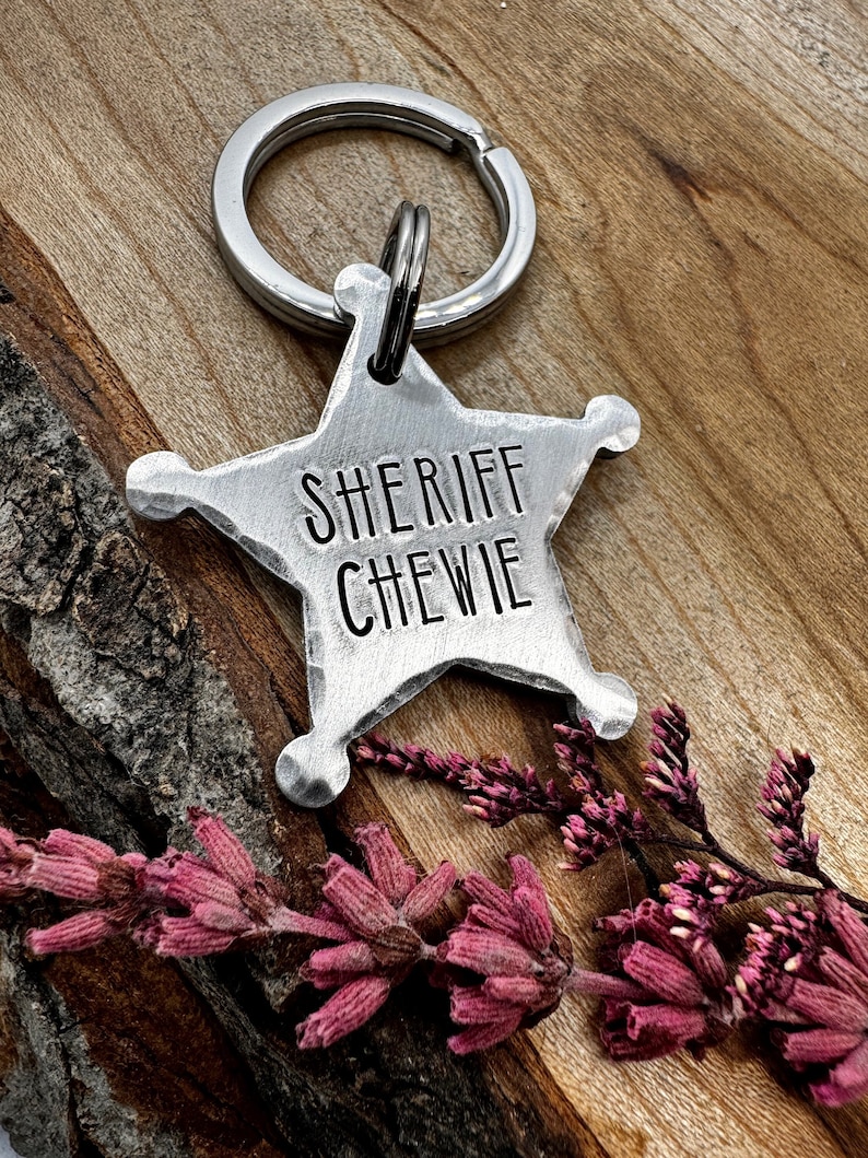 Op de afbeelding: Een zilveren sheriffster-sleutelhanger met de tekst "SHERIFF CHEWIE" erop gegraveerd. De sleutelhanger is bevestigd aan een zilveren sleutelring en ligt op een houten oppervlak met roze bloemen.
