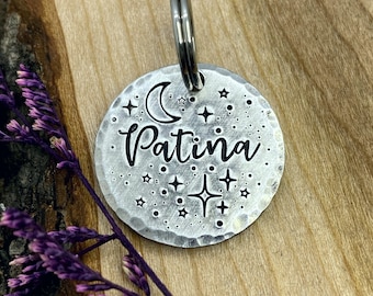 Puede incluir: Una etiqueta redonda para mascotas de color plateado con un acabado martillado. La etiqueta presenta una luna creciente, estrellas y la palabra "Patina" grabada en una fuente de escritura.