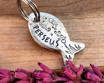 May include: Silver fish-shaped keychain with a ring, engraved with the name "PERSEUS" and a seahorse and jellyfish design.