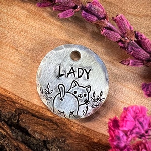 May include: A silver pet tag with the name "LADY" engraved on it. The cat is depicted in a playful pose with its tail up in the air. The tag has a small hole for attaching a leash or collar.