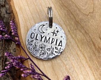 May include: Silver dog tag with the name "OLYMPIA" engraved on it. The tag is decorated with a crescent moon, stars, and floral designs.