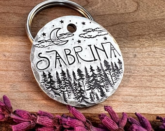 May include: Silver key chain with a round tag featuring a moon, stars, clouds, and a forest scene. The tag is engraved with the name "Sabrina".