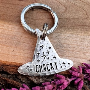 Pode incluir: Etiqueta de identificação para animais de estimação em prata em forma de chapéu de bruxa com o nome "CHUCKY" gravado. A etiqueta tem um chaveiro de prata e é decorada com pequenos recortes em forma de estrela.