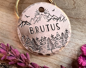 Könnte beinhalten: Ein kupferner Hundemarken mit einer Bergszene und dem eingravierten Namen "BRUTUS". Die Marke ist an einem goldfarbenen Schlüsselring befestigt.