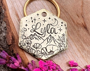May include: Gold-toned hexagonal pet tag with a mountain, moon, and flower design. The tag is engraved with the name "Lyla" and has a ring for attaching to a collar.