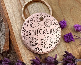 May include: A round, copper-colored metal pet identification tag featuring a hammered texture and engraved black outlines of chocolate chip cookies. The name SNICKERS is centered in black capital letters. A circular metal ring is attached at the top.