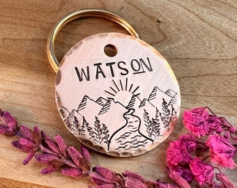 Könnte beinhalten: Ein runder, kupferfarbener Hundemarkenanhänger mit einer Bergszene und dem eingravierten Namen "WATSON". Der Anhänger ist an einem goldfarbenen Schlüsselring befestigt.