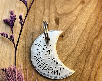 May include: A silver crescent moon-shaped pet tag with the name "SHADOW" engraved in a script font. The tag is decorated with a starburst and small stars.