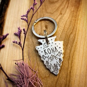Op de afbeelding: Zilveren hondenhanger in de vorm van een pijlpunt met een berglandschap, een halve maan en sterren. De hanger is gegraveerd met de naam "KONA" en bevestigd aan een zilveren sleutelhanger.