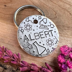 May include: Silver key chain with a round tag featuring the name "ALBERT" and science-themed illustrations, including a beaker, microscope, and atoms. The tag is attached to a silver key ring.