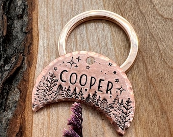 May include: Copper key chain with a crescent moon design featuring a forest silhouette and the name "COOPER" engraved in a script font. The key chain has a small ring for attaching keys.