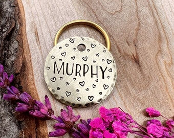 Könnte beinhalten: Ein runder, goldfarbener Hundemarkenanhänger mit einem Ring zum Anbringen an einem Halsband. Der Anhänger ist mit dem Namen "MURPHY" graviert und hat ein Muster aus kleinen Herzen am Rand.