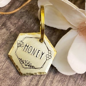 May include: A brass hexagon-shaped keyring with a hammered finish. The keyring features a bee, honeycomb, and floral design. The word "Honey" is engraved on the keyring.