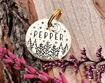 May include: A silver round pet tag with a black engraved design of a crescent moon, stars, and pine trees. The tag reads "PEPPER" and has a gold metal ring attached.