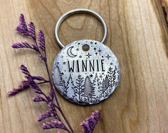 May include: Silver round pet tag with a moon, stars, and a forest design. The tag is engraved with the name "Winnie".