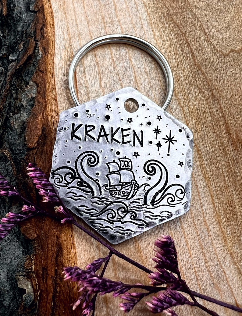 May include: A silver hexagonal key chain with a kraken and a ship engraved on it. The key chain has the word "KRAKEN" engraved on it. The key chain is on a wooden surface.