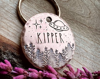 May include: A round copper pet tag with a hammered texture, featuring an engraved UFO, stars, and a forest of pine trees. The name KIPPER is stamped in the center. The tag is attached to a gold-toned metal split ring for easy attachment to a collar.