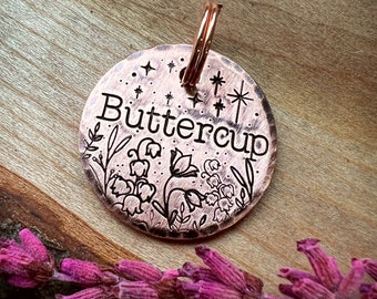 May include: A copper-colored pet tag with a floral design and the name "Buttercup" engraved on it. The tag has a ring attached to it.
