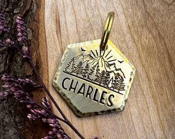 Könnte beinhalten: Ein sechseckiger Hundeanhänger aus Messing mit einer Bergszene und dem eingravierten Namen "CHARLES". Der Anhänger hat einen Ring zum Anbringen an einem Halsband.