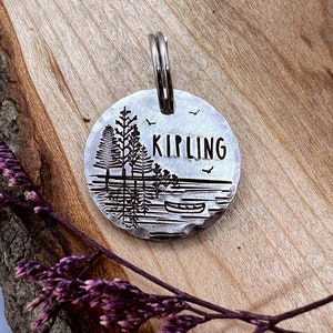 Könnte beinhalten: Silberner Hundeanhänger mit einer Silhouette von Bäumen und einem Kanu auf einem See. Der Anhänger ist mit dem Namen "KIPLING" graviert.