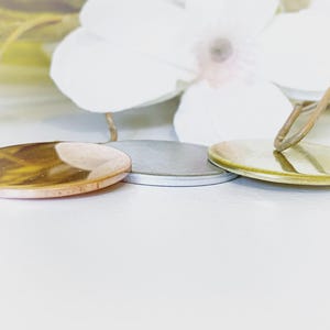 May include: Three circular metal pendants arranged in a row. The left pendant has a polished copper finish, the center pendant features a matte silver tone, and the right pendant is finished in a bright gold tone with a small metal loop attached.