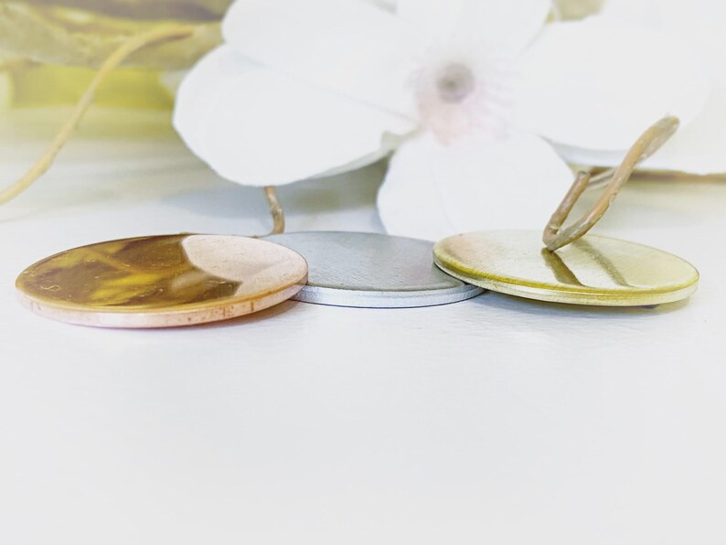 May include: Three circular metal pendants arranged in a row. The left pendant has a polished copper finish, the center pendant features a matte silver tone, and the right pendant is finished in a bright gold tone with a small metal loop attached.